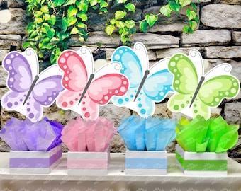 May include: Four butterfly centerpieces in purple, pink, blue, and green. Each centerpiece features a butterfly cutout with polka dots and a tissue paper base in a matching color. The centerpieces are displayed on a white shelf against a stone wall background.