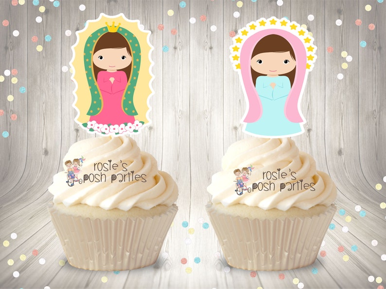 May include: Two cupcakes with white frosting topped with edible images of the Virgin Mary. The images are on white cardstock with a scalloped edge. The cupcakes are on a woodgrain background.