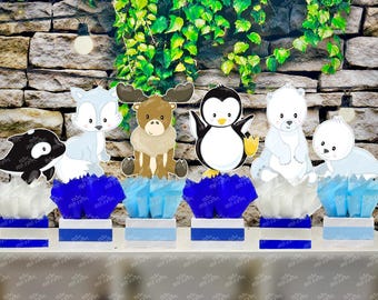 May include: A collection of animal-themed party decorations. The decorations include a black and white orca, a light blue fox, a brown moose, a penguin, a polar bear, and a seal. Each animal sits atop a blue and white box with tissue paper.