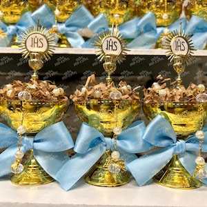 May include: Gold chalice-shaped containers with blue bows and a small gold cross with the letters "JHS" on top. The containers are filled with brown shredded paper and white beads.