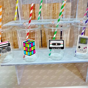 May include: Six clear plastic cups with colorful straws and retro themed stickers. The stickers feature a cassette tape, a game console, a boombox, and a Rubik's cube.