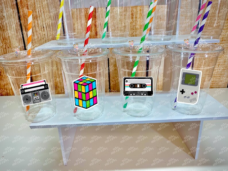 May include: Six clear plastic cups with colorful straws and retro themed stickers. The stickers feature a cassette tape, a game console, a boombox, and a Rubik's cube.
