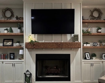May include: A white fireplace with a black mantel and a black fireplace screen. The fireplace is flanked by white cabinets with black hardware. There are wooden shelves above the cabinets, decorated with plants, wreaths, and framed pictures.