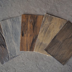 May include: Five wood grain samples in different shades of brown and gray. The samples are arranged in a fan shape on a gray stone surface.