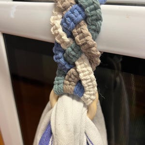 May include: A braided macrame towel holder in shades of blue, green, beige, and white, attached to a white appliance handle. A white and blue striped towel hangs from a wooden ring.