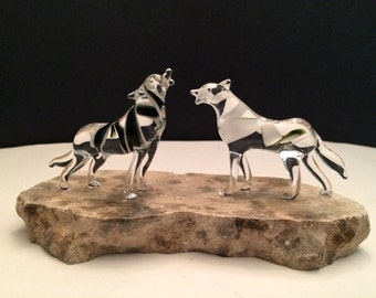 May include: Two clear glass wolf figurines howling at each other, standing on a small, rough, gray stone base.