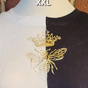May include: XXL two-tone t-shirt, split vertically with white and black panels. A gold-coloured bee and crown design is centered on the chest. The shirt is displayed on a mannequin.