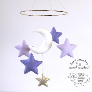 Può includere: Una luna crescente di feltro bianco e cinque stelle di feltro sono appese a un cerchio di metallo dorato. Quattro delle stelle sono viola e una è dorata. Il testo "miglior qualità cucito a mano" e "100% lana merino" è visibile nell'immagine.