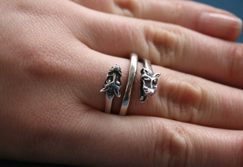 May include: A silver ring with two animal heads on it. The ring is wrapped around a finger.