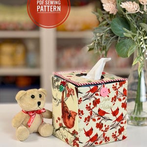 May include: A decorative tissue box cover featuring a red and cream cardinal and floral pattern. The box has a red and white striped trim and a pink button. A small teddy bear with a red ribbon is next to the box. The text "PDF Sewing Pattern" is in a red circle.