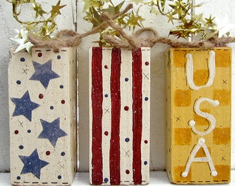May include: Three wooden blocks decorated with patriotic themes. The first block has blue stars on a white background with red dots. The second block has red and white stripes with blue dots. The third block has a yellow and white checkered pattern with the letters "USA" in white.