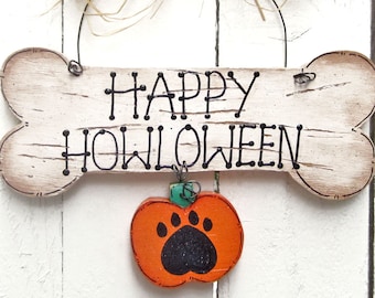Op de afbeelding: Een houten bord in de vorm van een bot met de tekst "Happy Howl-oween" in zwarte letters. Een kleine oranje pompoen met een zwarte pootprint hangt aan het bord.