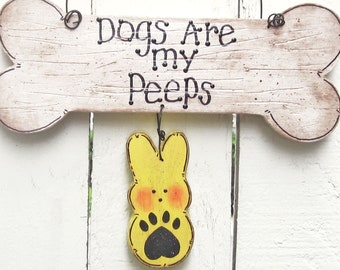 Op de afbeelding: Een houten bord in de vorm van een bot met de tekst "Dogs Are My Peeps" hangend aan een draad met een raffia strik. Een geel geverfd houten kuiken met een zwarte poot afdruk hangt aan het bord.