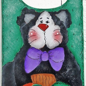 May include: A green wooden sign with the words "Welcome Fall" painted in black. A black cat with a purple bow tie is holding a red pumpkin with green leaves.