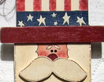 Op de afbeelding: Een houten welkomstbord met een rood, wit en blauw Amerikaans vlagontwerp. Het bord heeft een geschilderd gezicht met een snor en het woord "WELCOME" in witte letters op een blauwe achtergrond.