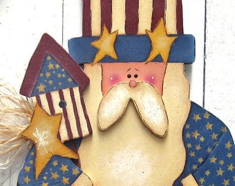 May include: A wooden folk art figurine of a patriotic Uncle Sam holding a star and a small American flag. He is wearing a blue and red striped hat, a blue and white star-patterned shirt, and red and white striped pants. The star he is holding says "Liberty to All".