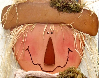 Peut inclure: Un visage de scarecrow en bois avec un chapeau de paille, un nez brun et un sourire. Le scarecrow est décoré de mousse et d'une écharpe en tissu blanc.