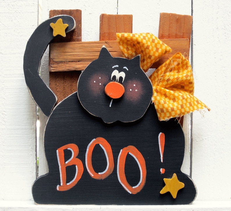 Puede incluir: Decoraci&oacute;n de Halloween de madera negra de un gato con un lazo amarillo y la palabra "BOO!" en letras naranjas. El gato tiene una nariz roja y ojos negros.