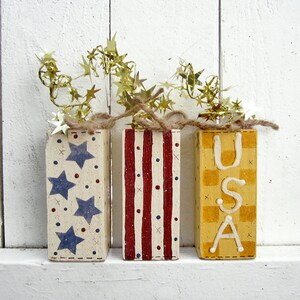 May include: Three wooden blocks painted in red, white, and blue with stars, stripes, and a checkered pattern. The blocks spell out "USA" and are decorated with gold star-shaped garland.