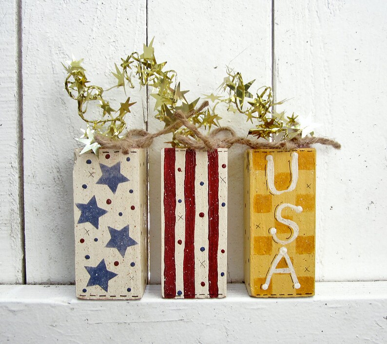 May include: Three wooden blocks painted in red, white, and blue with stars, stripes, and a checkered pattern. The blocks spell out "USA" and are decorated with gold star-shaped garland.