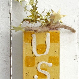 May include: A wooden block painted with a yellow and white checkered pattern. The letters "USA" are painted in white on the block. The block is decorated with a gold garland of stars.