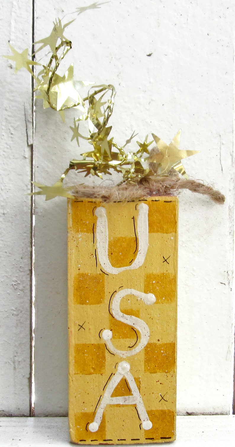 May include: A wooden block painted with a yellow and white checkered pattern. The letters "USA" are painted in white on the block. The block is decorated with a gold garland of stars.