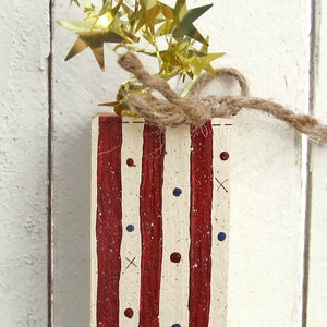 May include: A wooden block painted with red and white stripes and blue and red dots. The block is decorated with a gold star garland and twine.