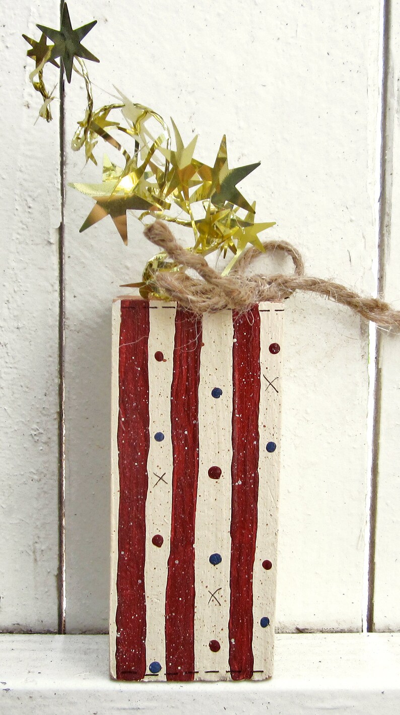 May include: A wooden block painted with red and white stripes and blue and red dots. The block is decorated with a gold star garland and twine.