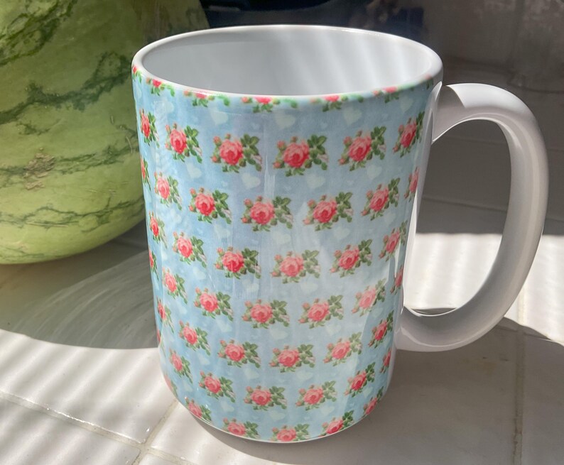 May include: A large ceramic mug with a white handle and interior. The exterior features a repeating pattern of pink roses on a light blue background. The mug is cylindrical and appears to be clean.