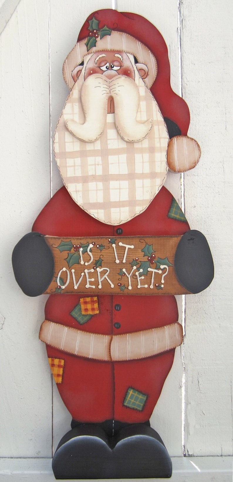 May include: A wooden Santa Claus figurine with a red hat, white beard, and plaid shirt. He is holding a sign that says "Is it over yet?"