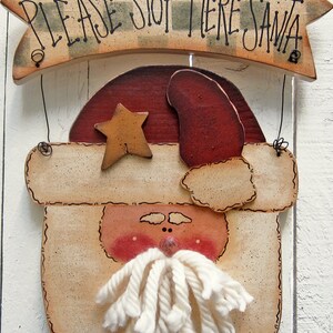 May include: A wooden sign with a painted Santa Claus face and the text "PLEASE STOP HERE SANTA" on a banner above. The Santa Claus face has a red hat, a white beard made of yarn, and a gold star on his hat.