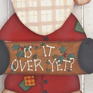 May include: A wooden Santa Claus figure with a red hat, white beard, and a sign that reads "Is it over yet?" The figure is wearing a red suit with green and yellow patches and black boots.