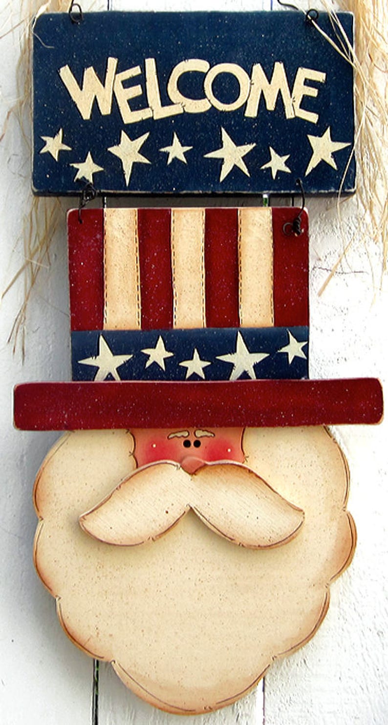 May include: Wooden welcome sign with a red, white, and blue American flag design. The sign features a white beard and a red hat with stars. The sign says "Welcome" in white letters.