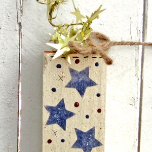 May include: A wooden block with a white background and blue stars painted on it. The block is decorated with red and blue dots and has a brown twine string attached to the top. The block is surrounded by gold star-shaped confetti.