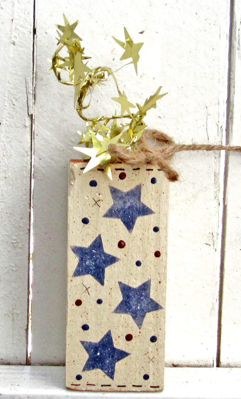 May include: A wooden block with a white background and blue stars painted on it. The block is decorated with red and blue dots and has a brown twine string attached to the top. The block is surrounded by gold star-shaped confetti.