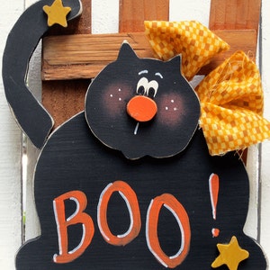 Puede incluir: Decoraci&oacute;n de Halloween de madera negra de un gato con un lazo amarillo y la palabra "BOO!" en letras naranjas. El gato tiene una nariz roja y ojos blancos.