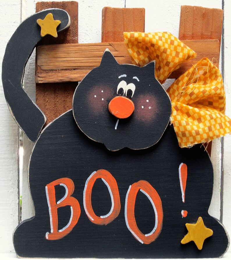 Puede incluir: Decoraci&oacute;n de Halloween de madera negra de un gato con un lazo amarillo y la palabra "BOO!" en letras naranjas. El gato tiene una nariz roja y ojos blancos.