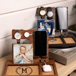 May include: A wooden phone charging station with a cork surface. It features a phone stand, a watch holder, a space for wireless charging, and a photo frame. The station is personalized with the letter "M" in a wreath.