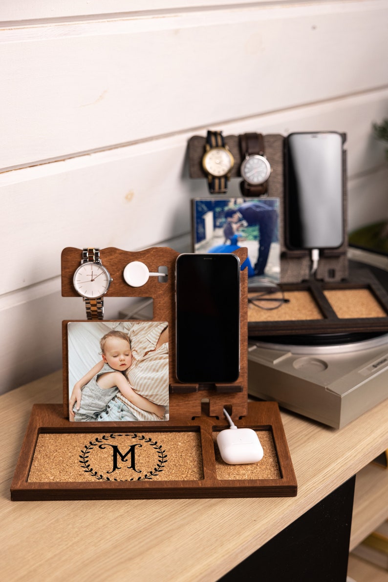 May include: A wooden phone charging station with a cork surface. It features a phone stand, a watch holder, a space for wireless charging, and a photo frame. The station is personalized with the letter "M" in a wreath.