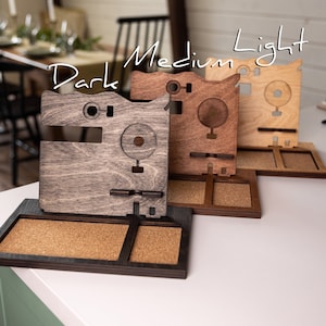 May include: Three wooden phone stands in different shades of brown, labeled "Dark", "Medium", and "Light". Each stand has a corkboard section and a cutout for a phone.