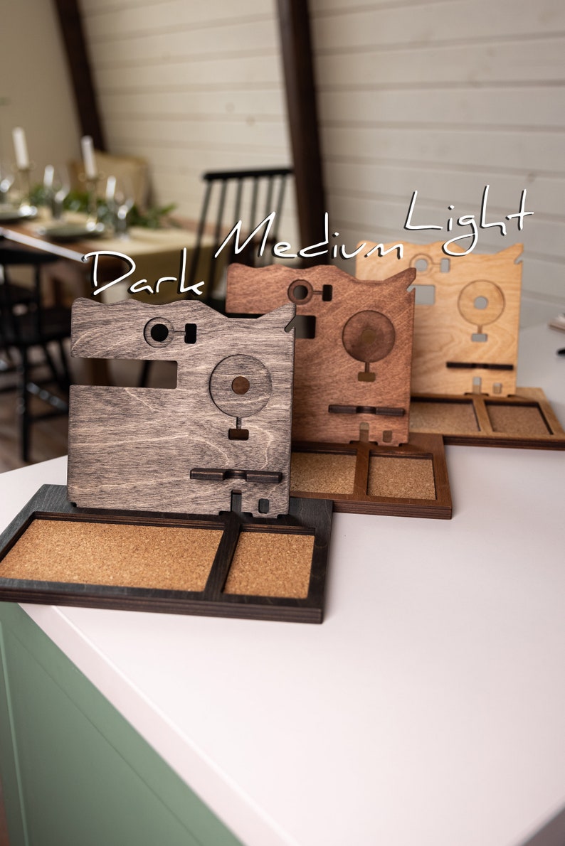 May include: Three wooden phone stands in different shades of brown, labeled "Dark", "Medium", and "Light". Each stand has a corkboard section and a cutout for a phone.