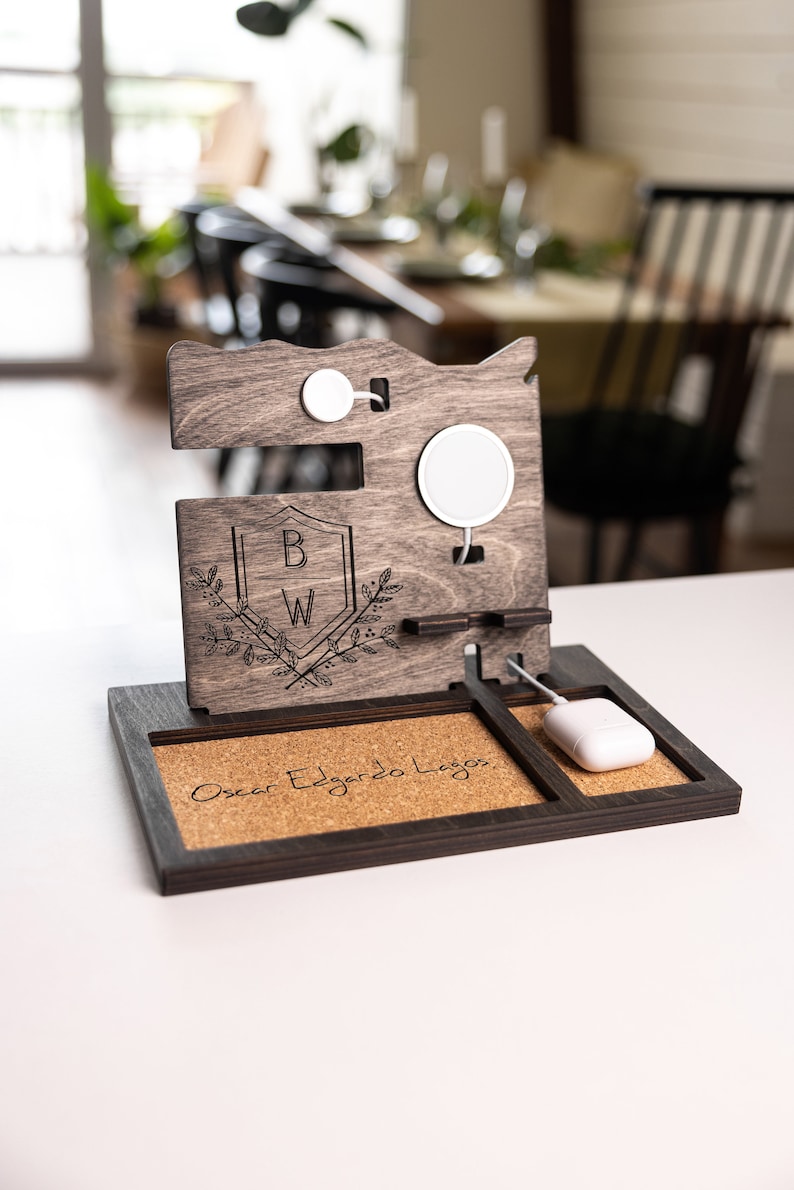May include: A wooden phone stand with a cork base. The stand has two charging pads, a watch holder, and a slot for a phone. The stand is personalized with the initials "B.W." and the name "Oscar Edgardo Lagos".