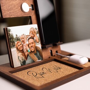 May include: A wooden phone stand with a cork insert. The stand has a slot for a phone, a space for wireless charging, and a small photo frame. The photo frame shows a family of four. The cork insert has the name "Ryan Walker" written on it.