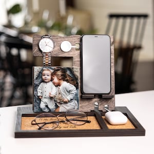 May include: Wooden phone stand with a cork base. It has a slot for a phone, a watch, a wireless charger, and a photo frame. The photo frame shows a picture of two children. The cork base has the text "To the Best Dad..." and "Love, Brady and Cole".