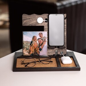 May include: A wooden phone stand with a cork base. The stand has a slot for a phone, a wireless charger, and a picture frame. The picture frame shows a family of four. The stand also has a slot for glasses and a small white wireless charging pad. The stand has the text "Dad you no matter what you are" on the cork base.