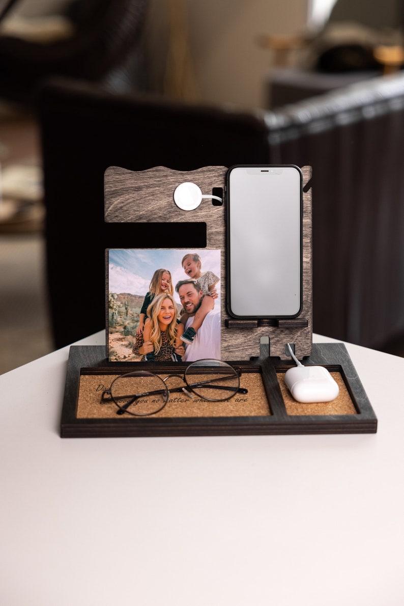 May include: A wooden phone stand with a cork base. The stand has a slot for a phone, a wireless charger, and a picture frame. The picture frame shows a family of four. The stand also has a slot for glasses and a small white wireless charging pad. The stand has the text "Dad you no matter what you are" on the cork base.