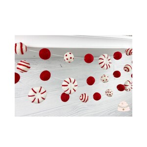 May include: A red and white felt garland with candy cane, polka dot, and striped ornaments. The garland is strung on a white string and is hanging from a white shelf.