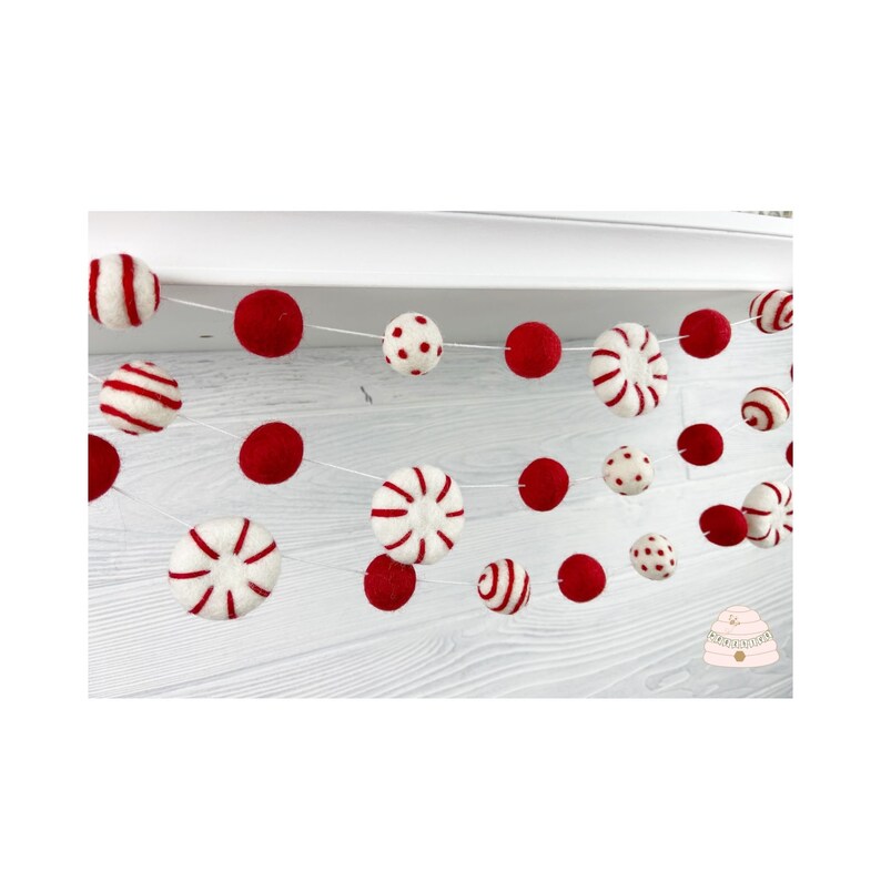 May include: A red and white felt garland with candy cane, polka dot, and striped ornaments. The garland is strung on a white string and is hanging from a white shelf.