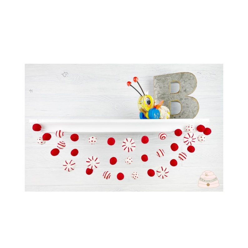 May include: A red and white peppermint garland with red pom poms hangs from a white shelf. The garland is made of felt and is perfect for a Christmas or winter themed party.
