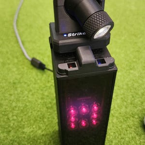 May include: A black StrikeLED device featuring a rectangular base with a vertical array of six glowing pink lights. A rotatable black flashlight head with a textured grip is mounted on top, displaying the text StrikeLED and Strike on the housing.